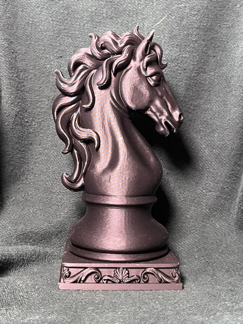A knight piece statue shaped like a horse head with a flowing, ornate mane, standing on a square scrollwork base against a charcoal green fabric backdrop.
