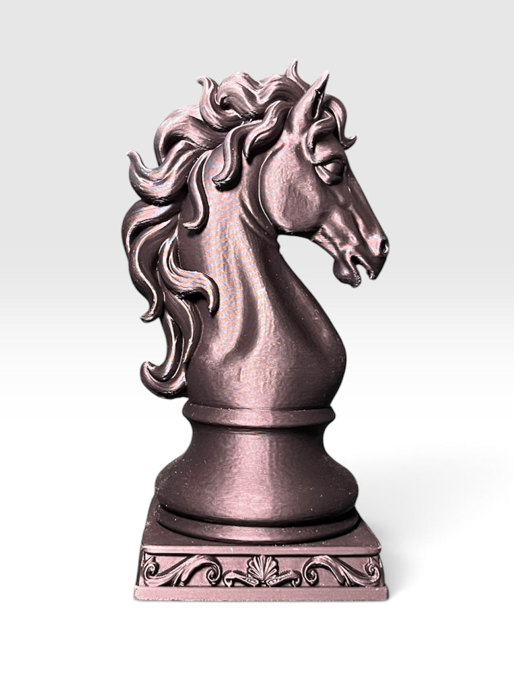 A knight chess piece statue shaped like a horse head with a flowing, ornate mane, standing on a square scrollwork base against a white to light grey gradient background.