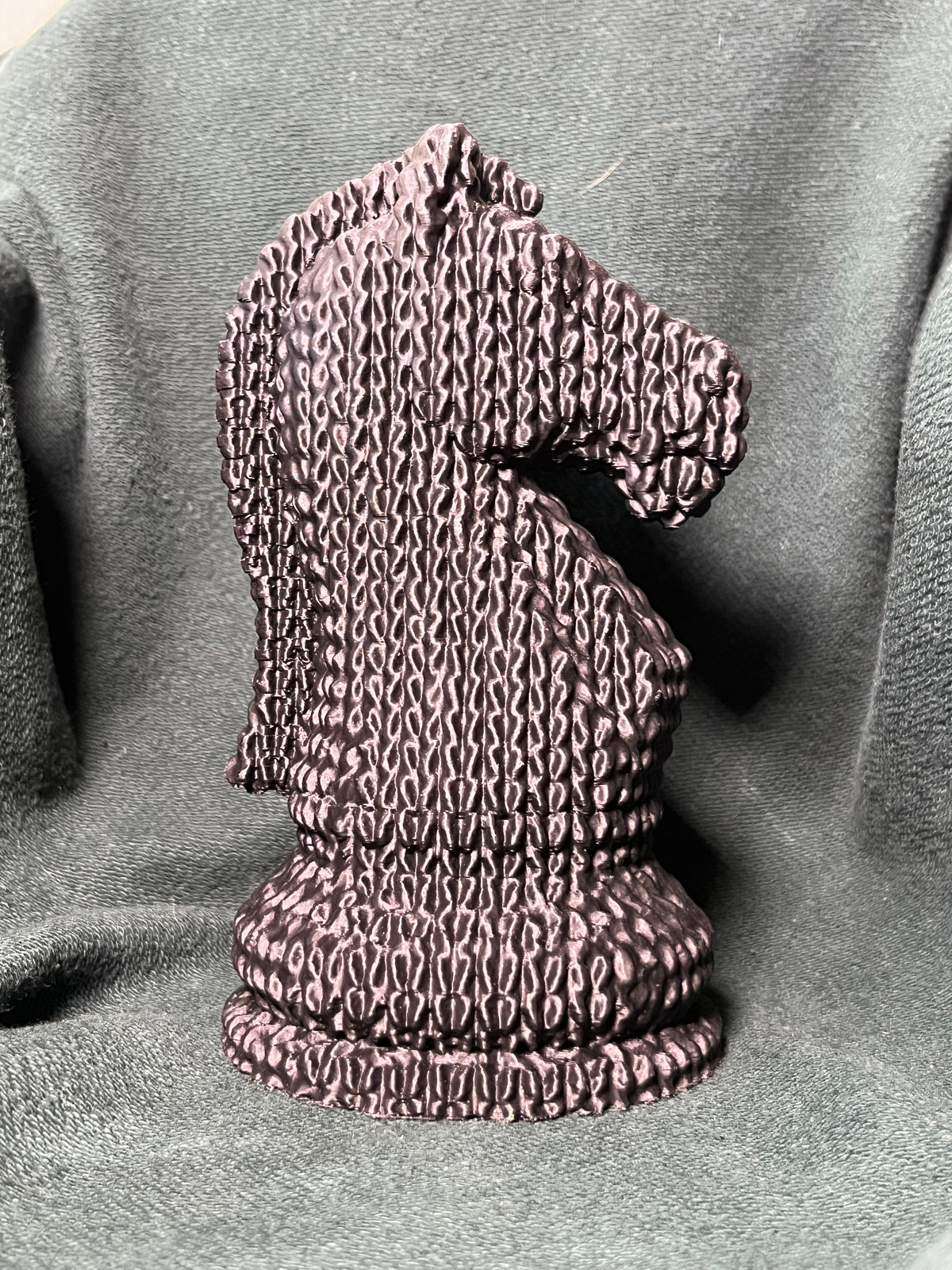 A knight piece statue shaped like a horse head sculpted with a knitted, stitched texture, standing on a round base against a charcoal green fabric backdrop.