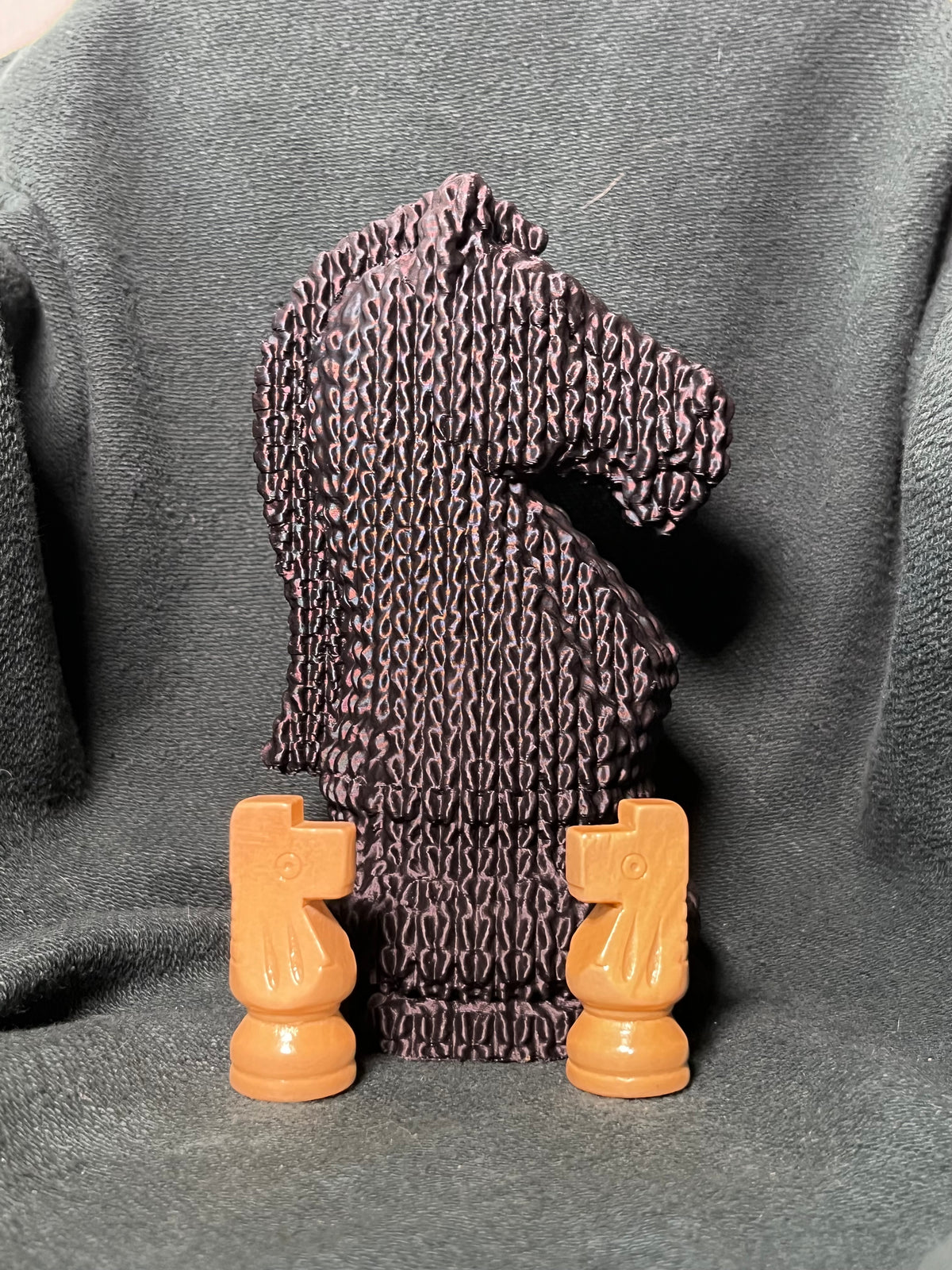 A jumbo knight chess piece statue sculpted with a knitted, stitched texture, photographed on a dark gray textured fabric background, with two tan knight chess pieces sitting in front for scale.