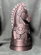 A knight piece statue shaped like a horse in mayan armor, standing on a round base against a charcoal green fabric backdrop.