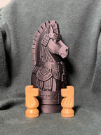 A molded giant chess piece statue with mayan armor, photographed on a dark gray textured fabric background. Two tan knight chess pieces sitting in front.