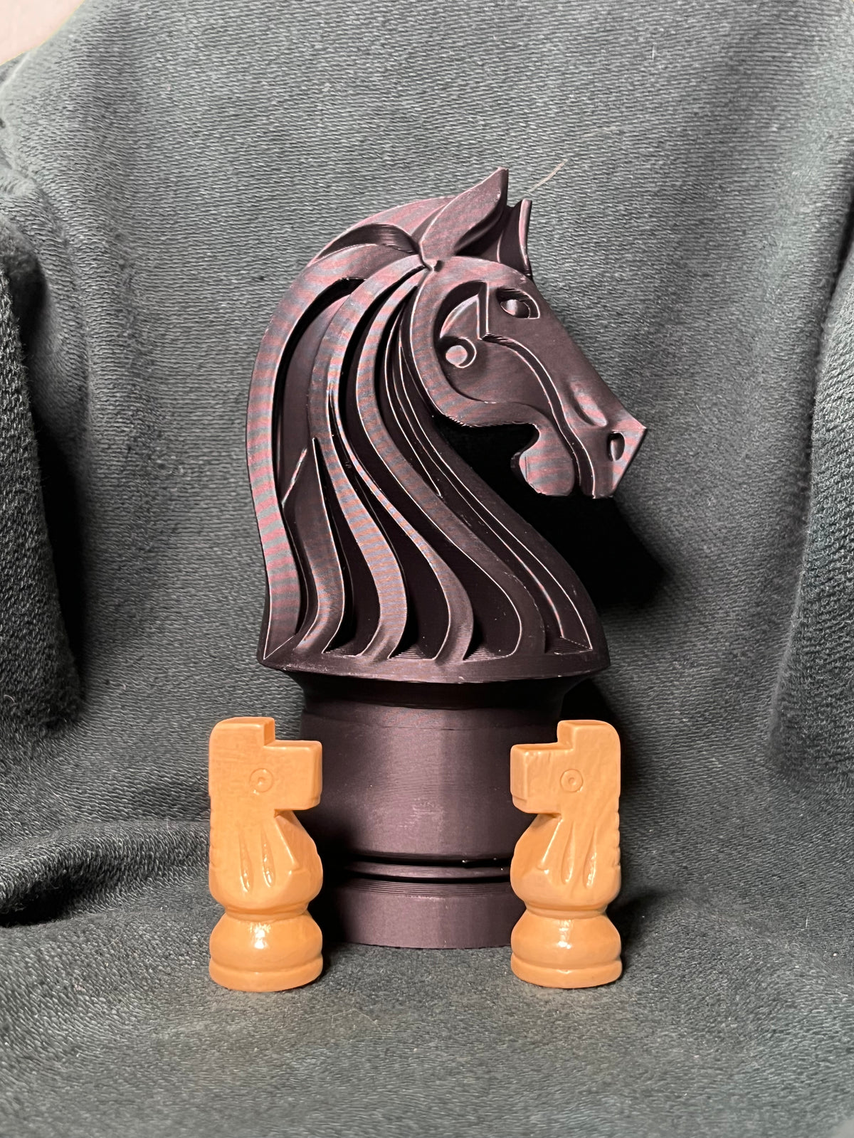 A jumbo knight chess piece statue with a flowing sculpted mane, photographed on a dark gray textured fabric background, with two tan knight chess pieces sitting in front for scale.